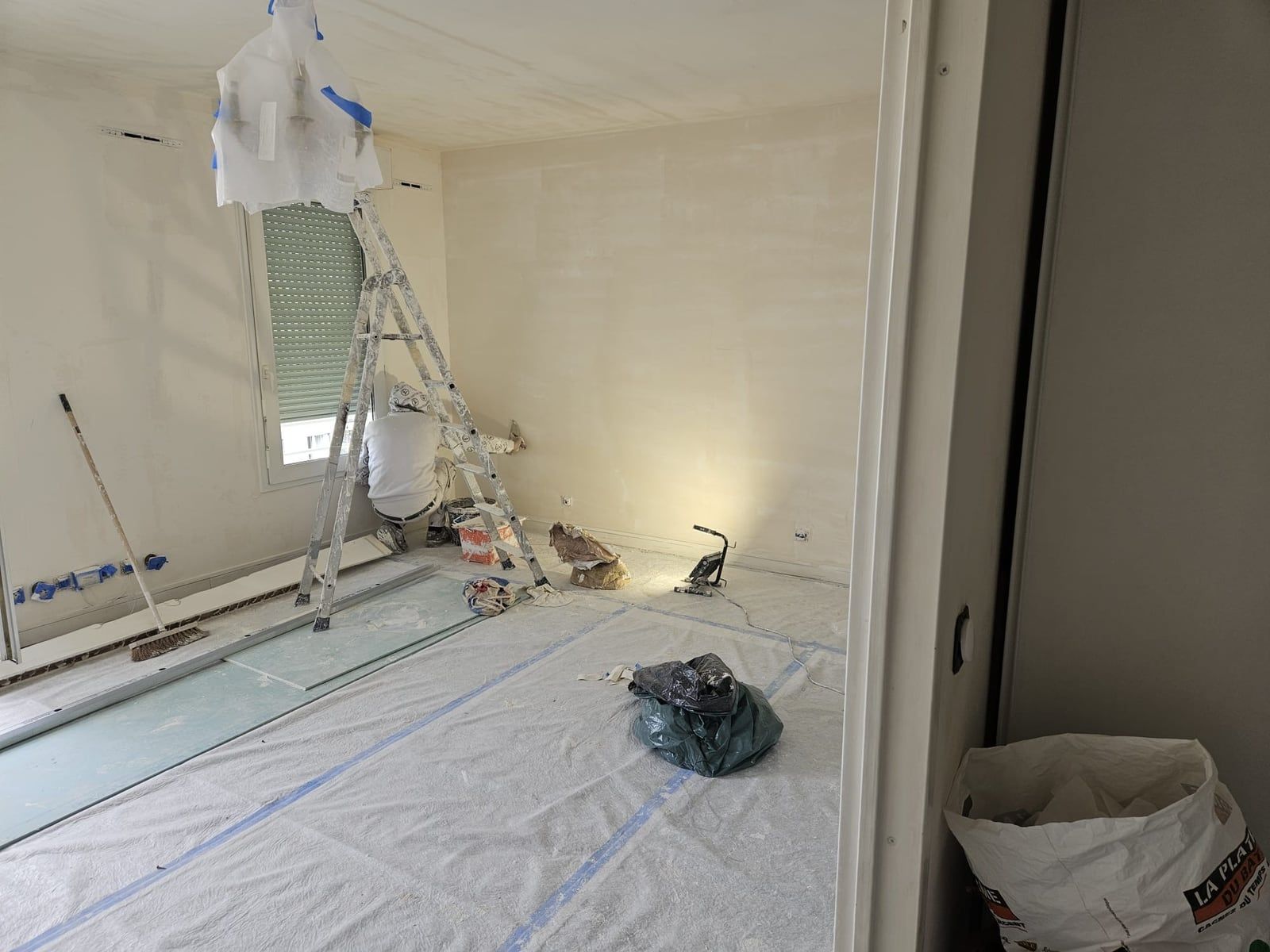 Séjour en chantier : sol entièrement bâché, escabeau et sacs de plâtre, murs en cours de finition